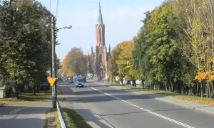 Jesień na drodze krajowej 91. W oddali widać kościół z wieżą, a po bokach drogi rosną drzewa w jesiennych barwach.