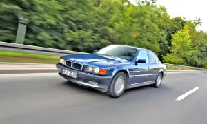 Błękitne BMW 750i pędzi po drodze, otoczone zielenią drzew.