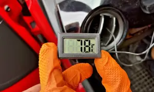 Rękawiczka w kolorze pomarańczowym trzyma termometr pokazujący 78.1 stopnia. Obok widać kratkę nawiewu, prawdopodobnie z samochodu z włączonym a/c.