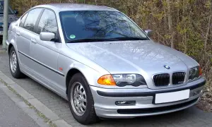 Srebrne BMW E46 330i stoi zaparkowane przy chodniku, otoczone zielenią.