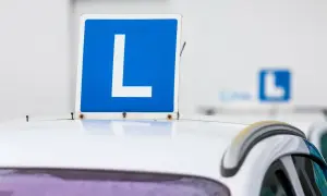Tablica "L" na dachu białego samochodu. Zbliża się egzamin na prawo jazdy, a ten znak symbolizuje naukę jazdy.