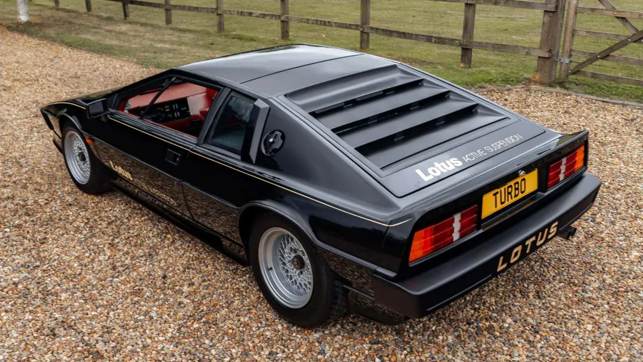 Czarny Lotus Esprit Turbo z czerwonym wnętrzem, zaparkowany na żwirze, z charakterystycznymi żaluzjami na tylnej szybie.