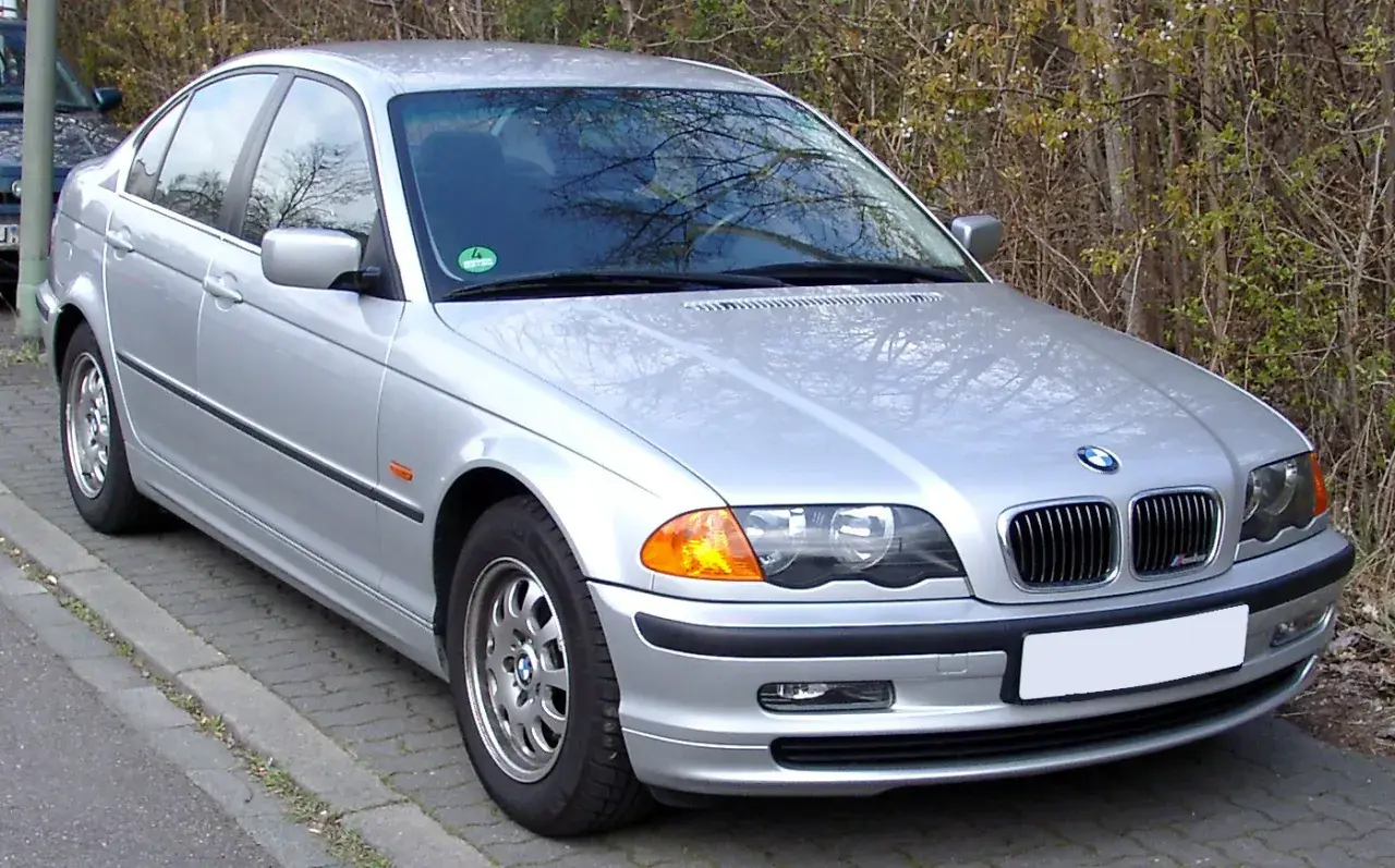 Srebrne BMW E46 330i stoi zaparkowane przy chodniku, otoczone zielenią.