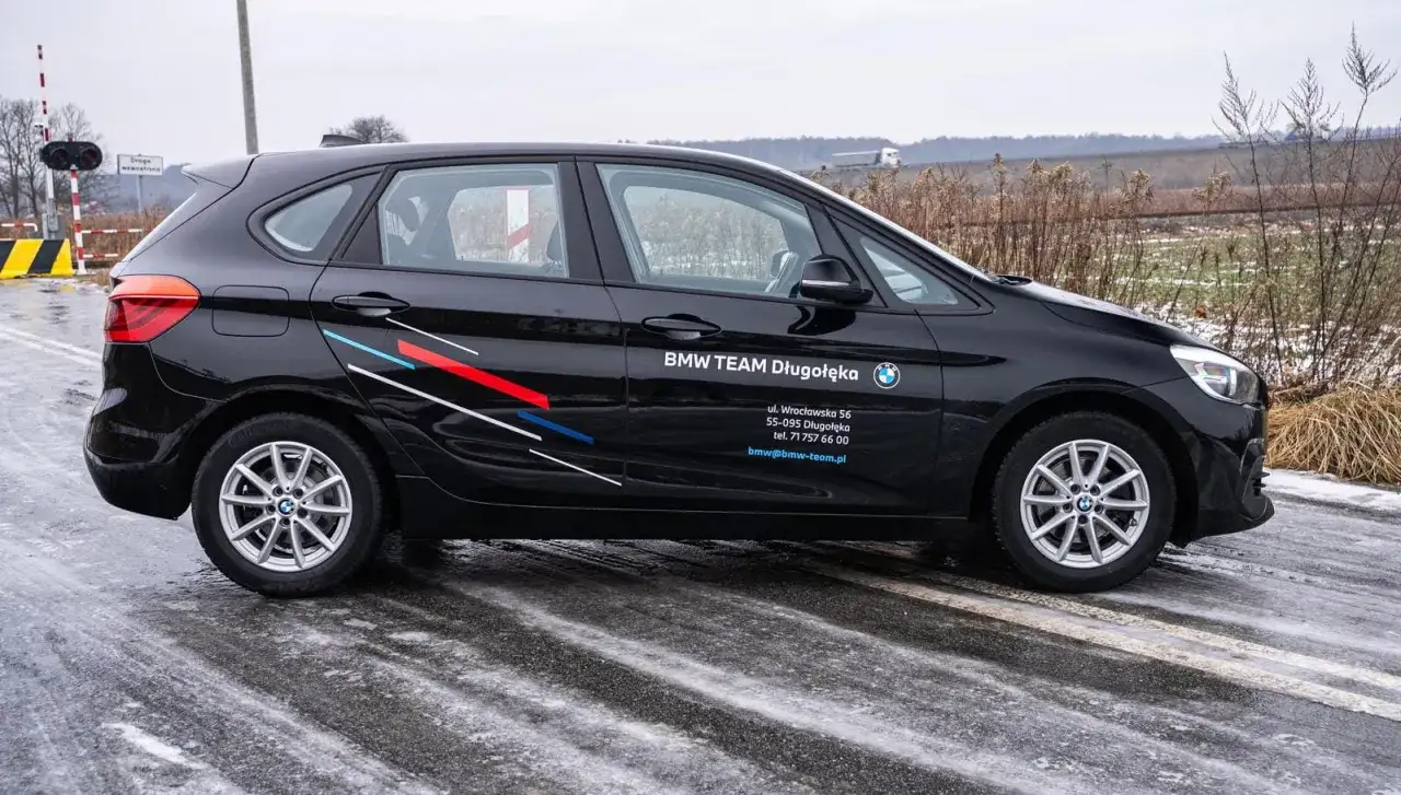 Czarne BMW F45 z logo BMW TEAM Długołęka stoi na mokrej drodze. W tle przejazd kolejowy i pola.