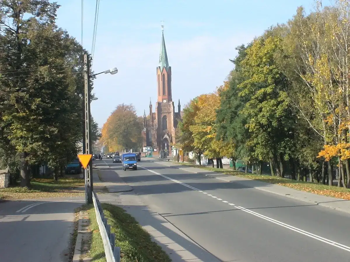 Jesień na drodze krajowej 91. W oddali widać kościół z wieżą, a po bokach drogi rosną drzewa w jesiennych barwach.