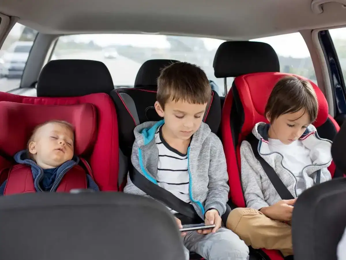 Maluch śpi w foteliku, starsi bracia grają na telefonach. Od kiedy dziecko może jeździć bez fotelika, zależy od jego wzrostu i wagi.