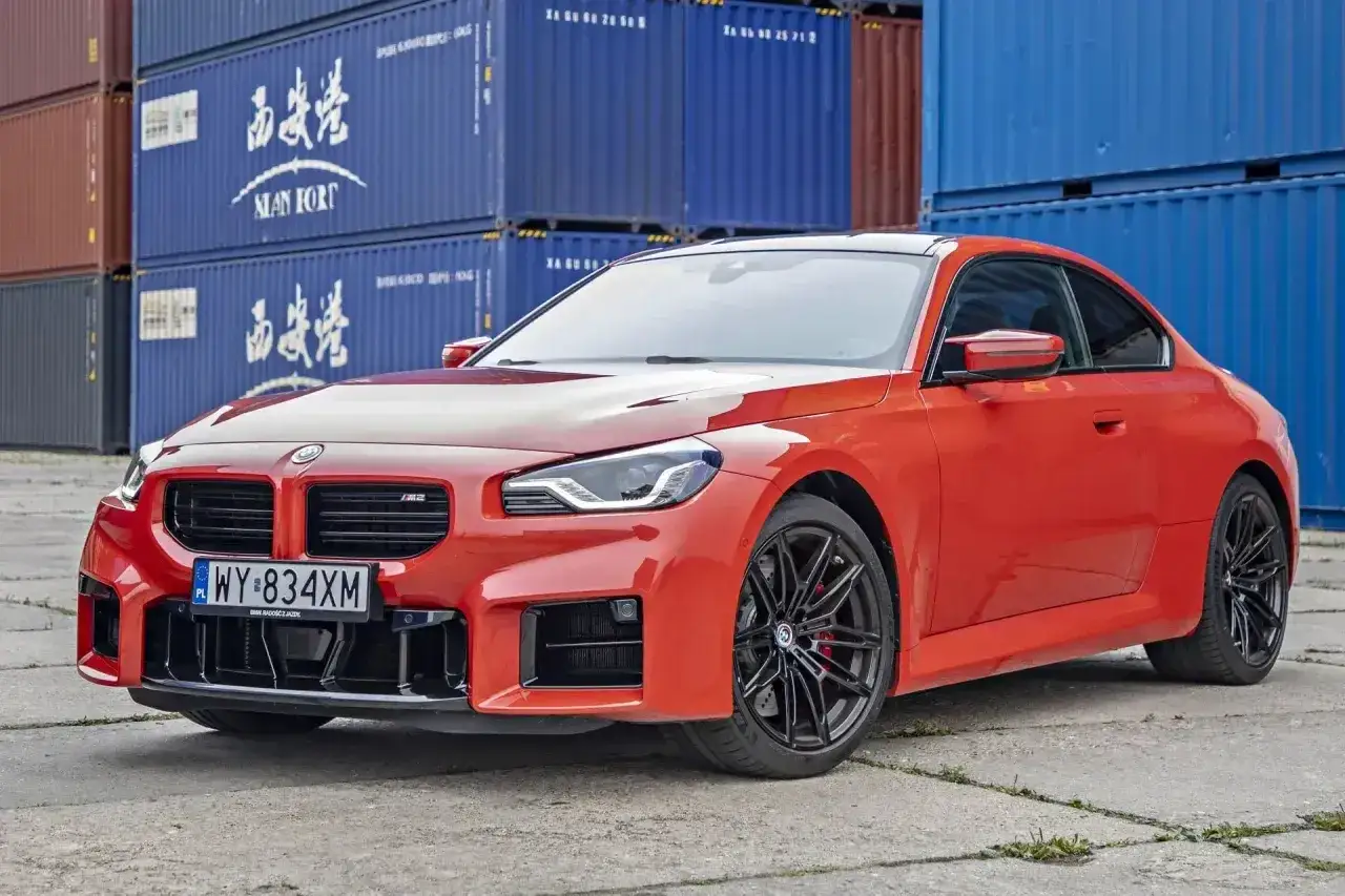 Pomarańczowe BMW M2 z czarnymi felgami, stojące przed niebieskimi kontenerami. Sprawdź aktualną bmw m2 cena.
