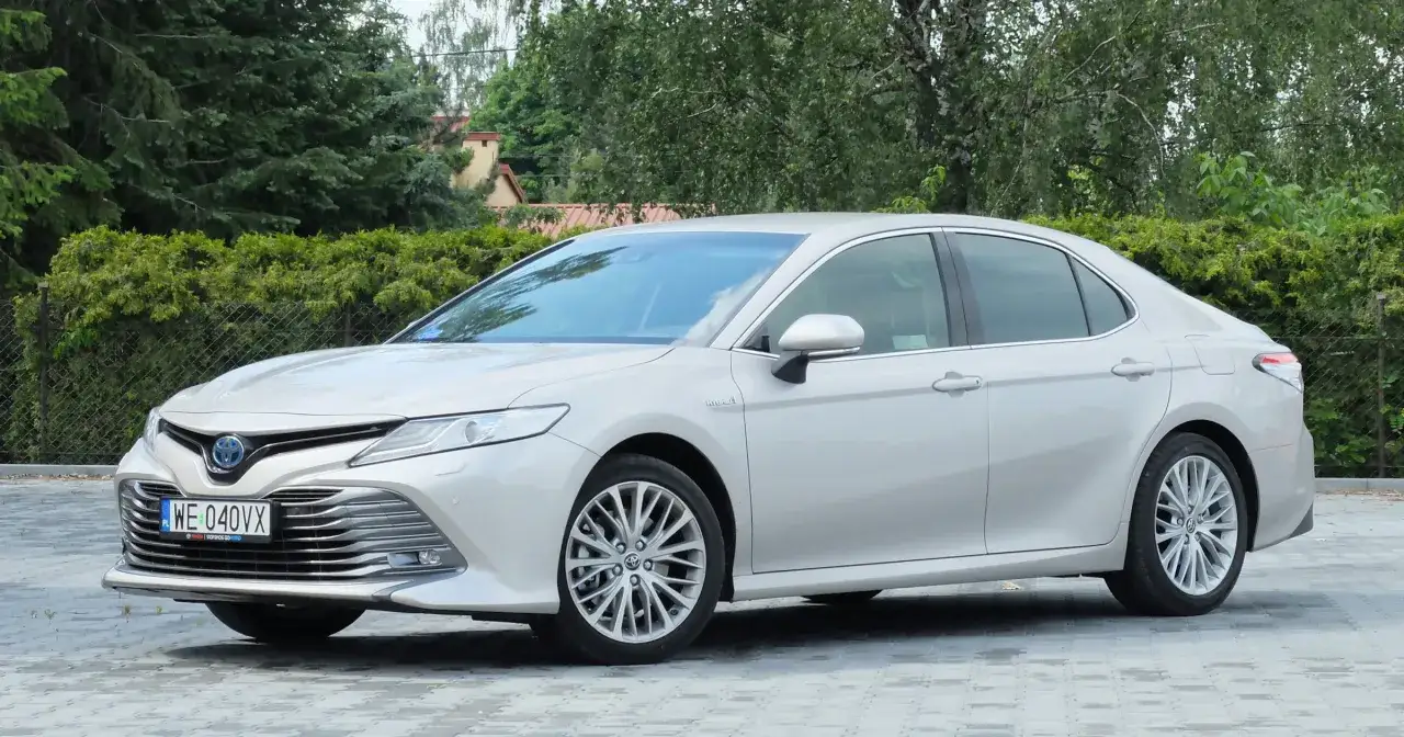 Elegancka, srebrna Toyota Camry 2.5 Hybrid stoi na wybrukowanym podjeździe, otoczona zielenią.