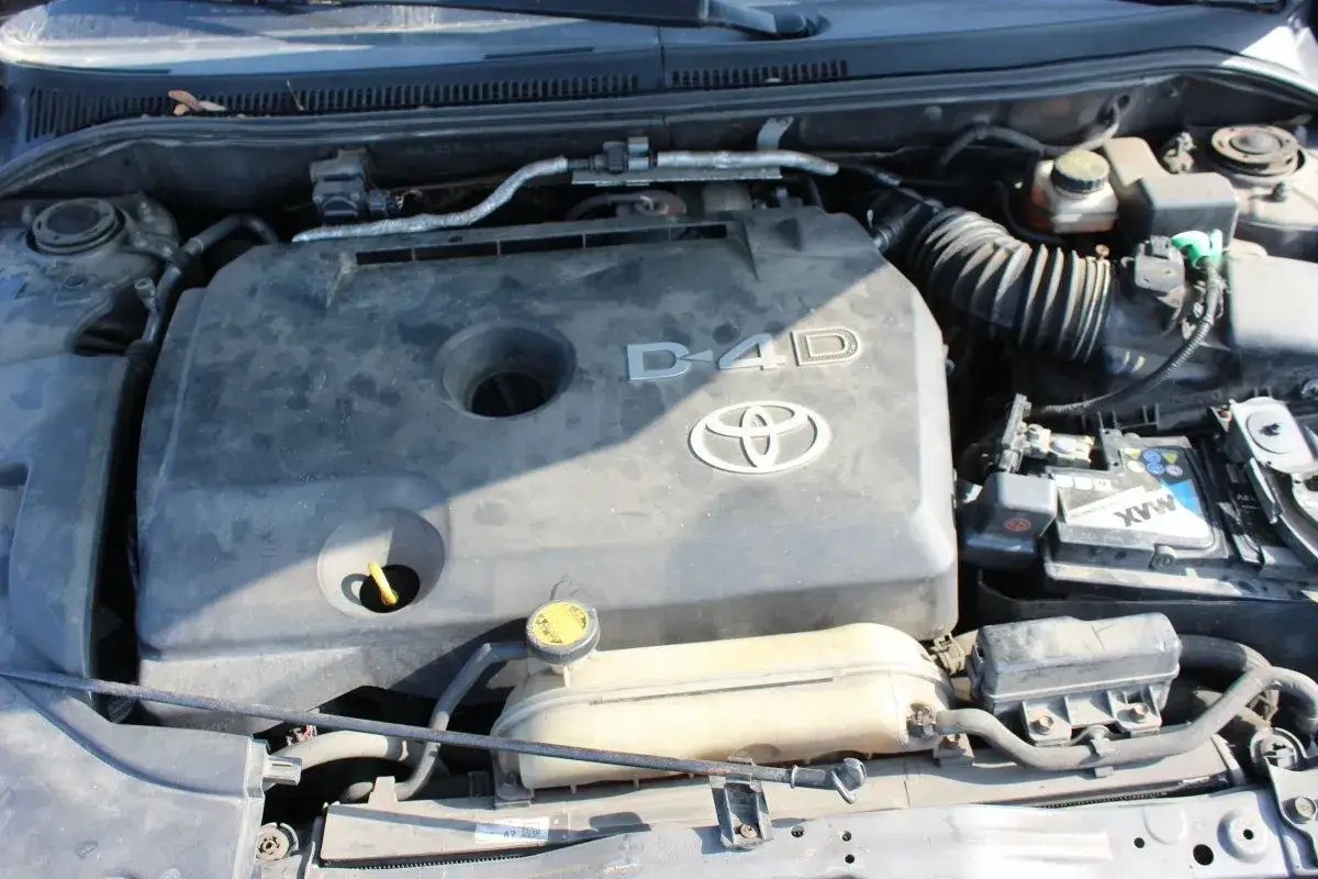 Silnik D-4D w Toyota Avensis 2.0. Widoczny jest plastikowy dekiel silnika z logo Toyoty i oznaczeniem D-4D, zbiornik płynu chłodniczego i akumulator.