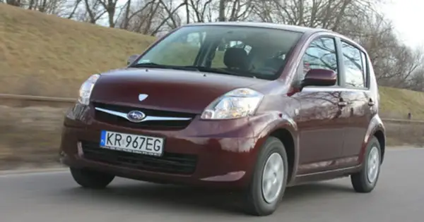 Ciemnoczerwony Subaru Justy jedzie po drodze, zalesione wzgórze w tle.