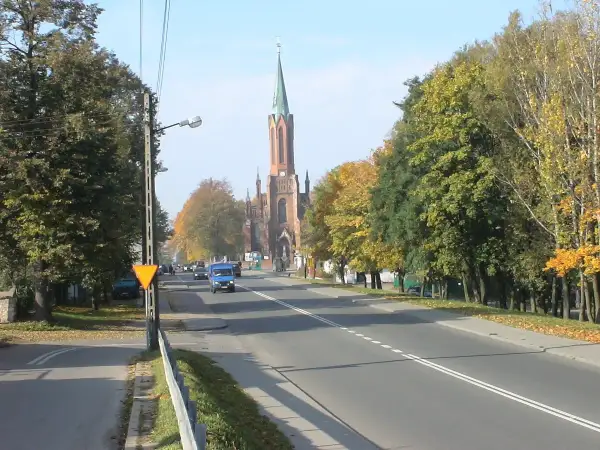 Jesień na drodze krajowej 91. W oddali widać kościół z wieżą, a po bokach drogi rosną drzewa w jesiennych barwach.