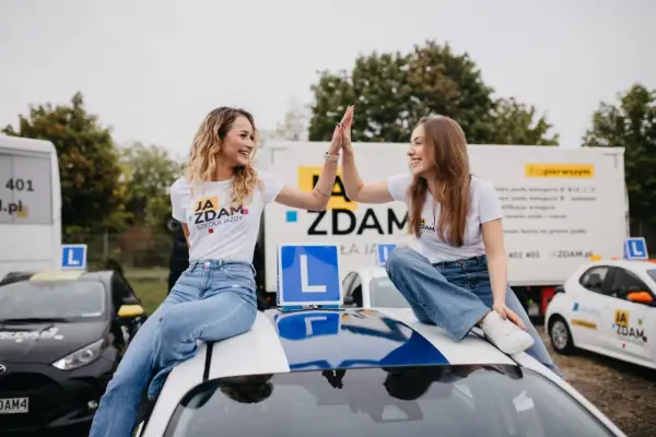 Dwie uśmiechnięte dziewczyny świętują zdanie egzaminu na prawo jazdy kat. B, siedząc na dachu samochodu szkoleniowego.