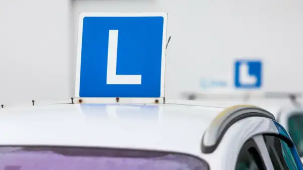 Tablica "L" na dachu białego samochodu. Zbliża się egzamin na prawo jazdy, a ten znak symbolizuje naukę jazdy.