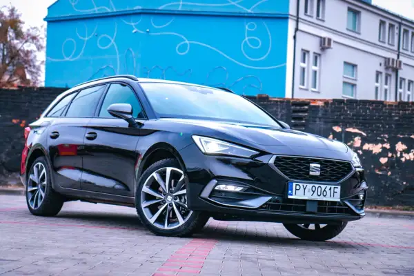 Czarny Cupra kombi z polskimi tablicami rejestracyjnymi stoi na brukowanym placu przed budynkiem z niebieską, ozdobną ścianą.