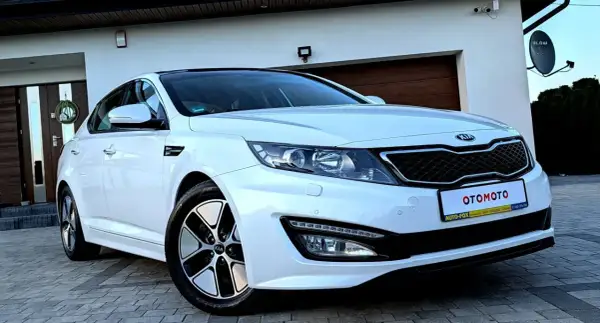 Biały kia sedan z czarnym dachem, zaparkowany przed domem z drewnianą bramą garażową.